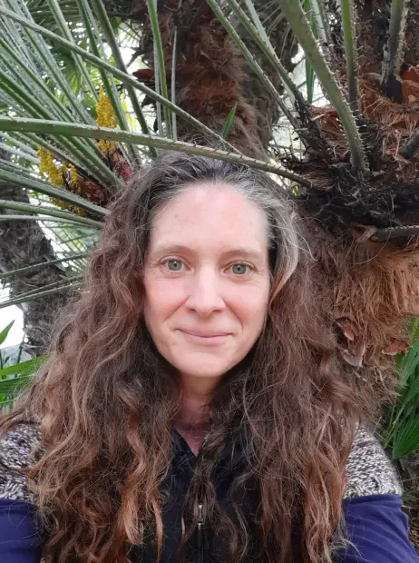 Woman with long curly hair in front of tall green plants, looking at the camera.