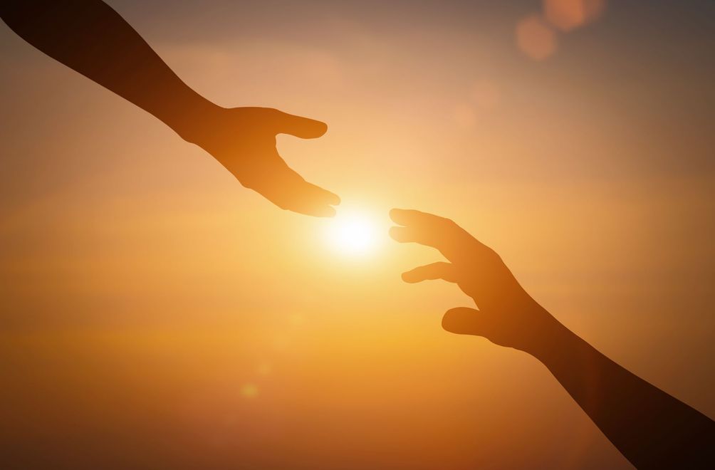 Two silhouetted hands reaching toward each other at sunset, warm orange sky glowing between them