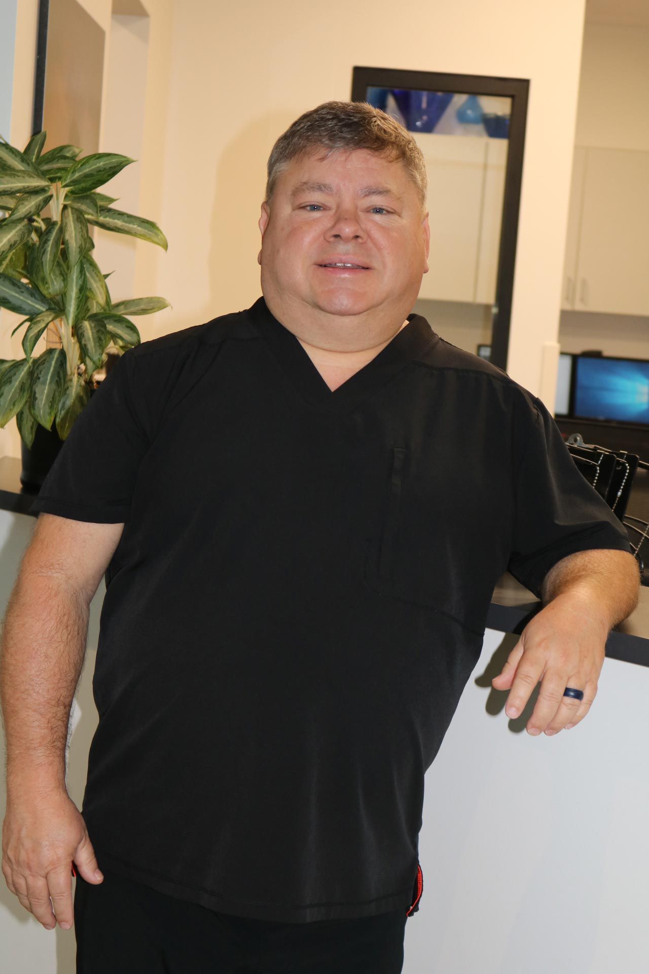 Man in black scrubs leans on a counter, smiling. Indoor setting with a plant and mirror. Man in black scrubs leans on a counter, smiling. Indoor setting with a plant and mirror.
