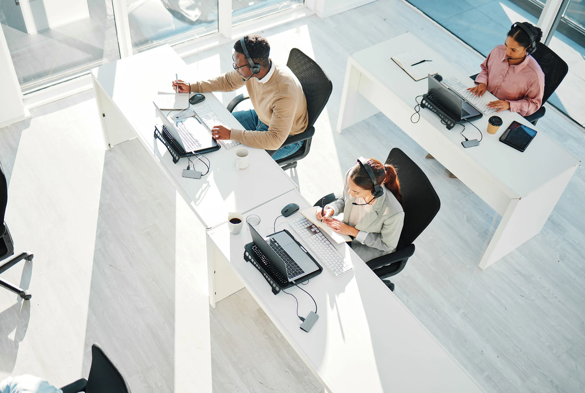 Three people in an office wearing headsets and working on laptops at white desks. Sunlight streams in.