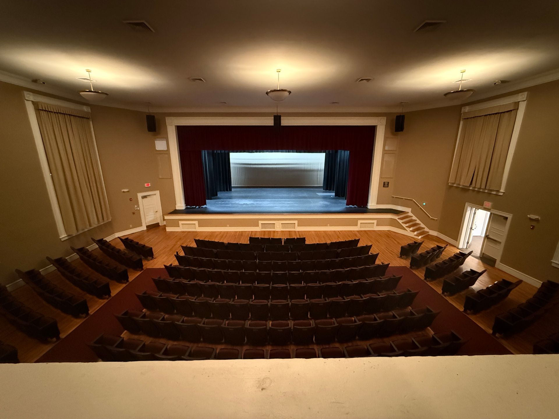 Auditorium with rows of seats facing a stage with a red curtain. Beige walls, wood floor.