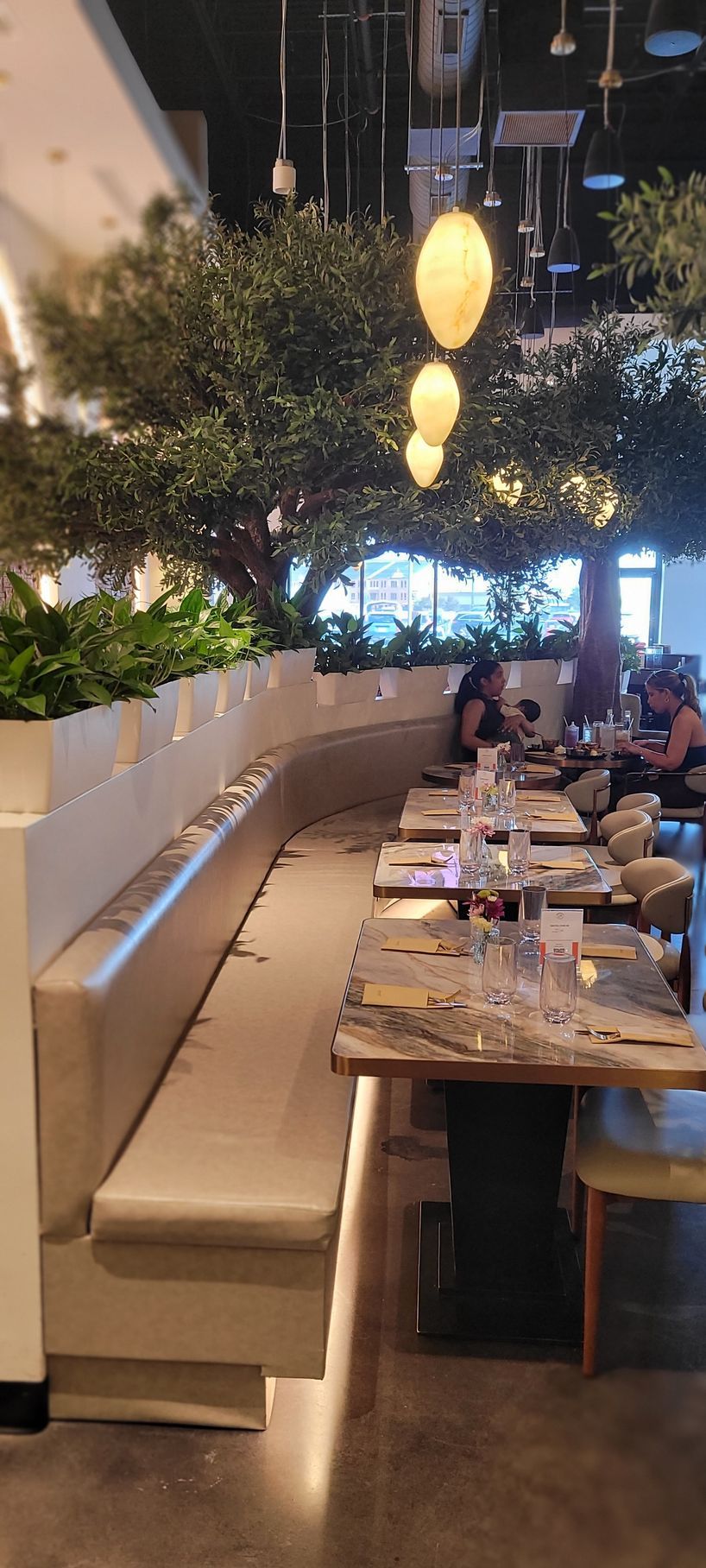 Restaurant dining area with a long table, white booth seating, hanging lights, and greenery decor