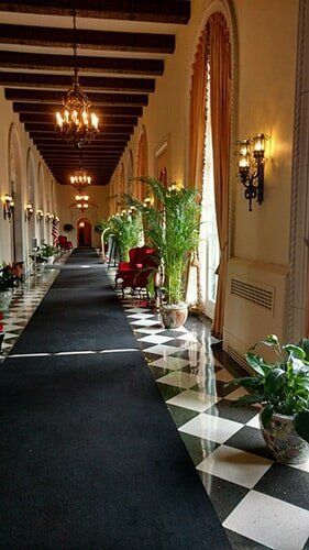 Long hallway with chandeliers, patterned tile, black runner, plants, and wall sconces
