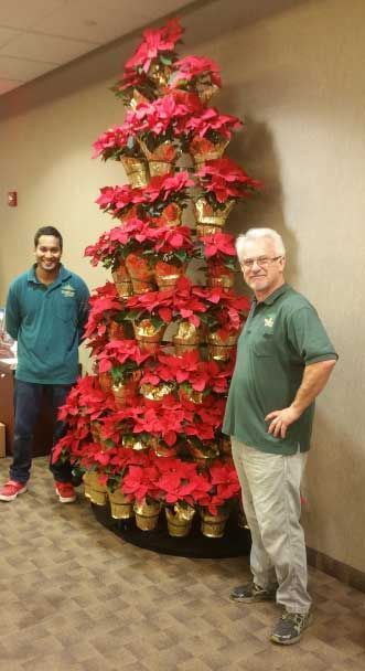 Two men pose beside a tall poinsettia display in a hallway.