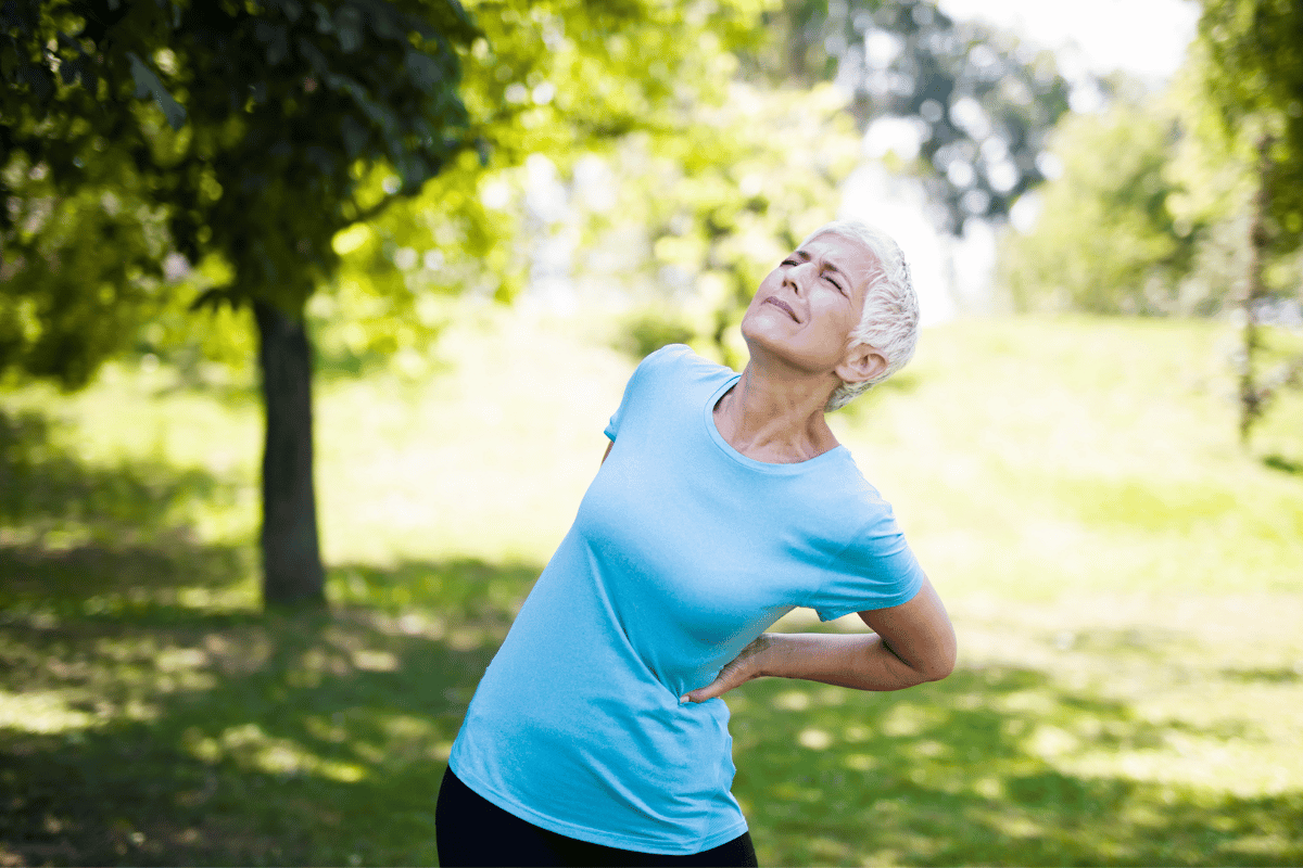 Woman with short gray hair, in blue shirt, clutches her lower back outdoors, in pain.