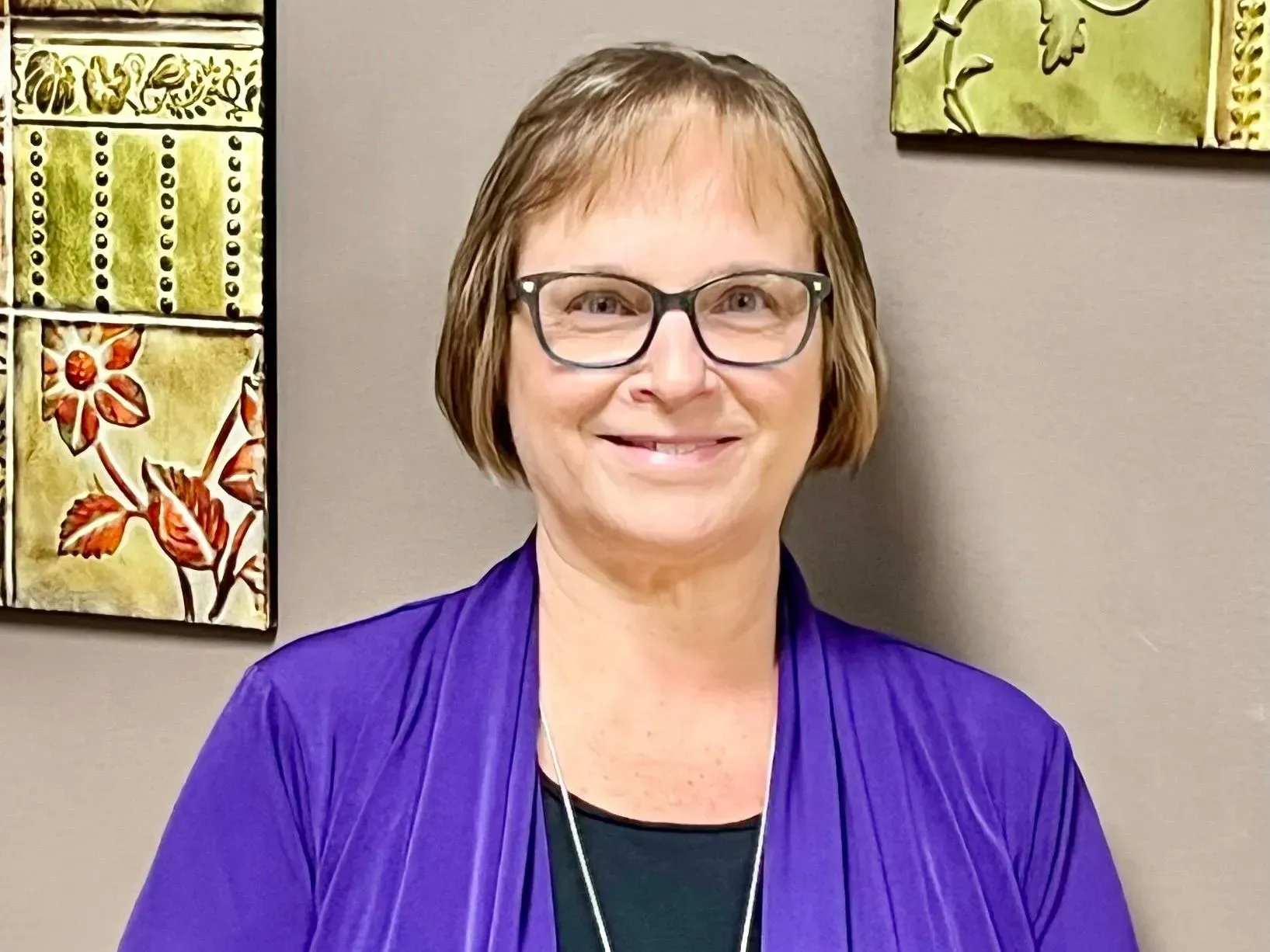 Woman with glasses smiling, wearing a purple top, standing in front of decorative wall tiles.