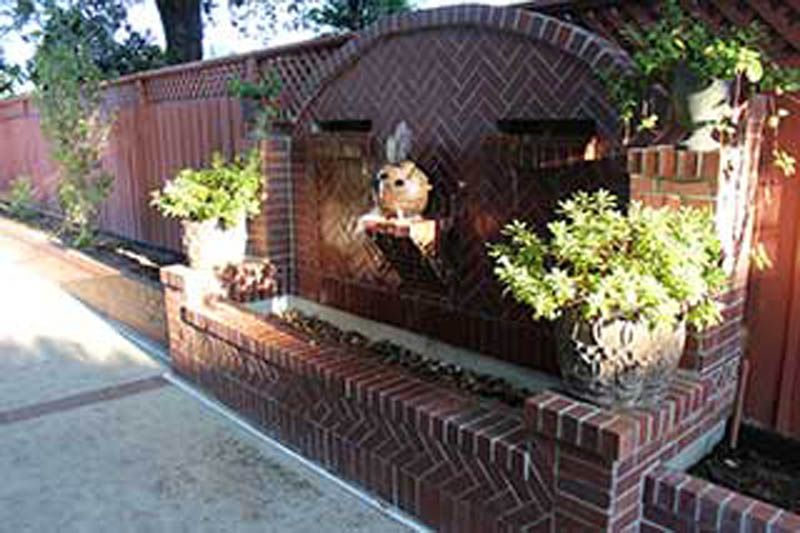 A brick wall with a waterfall and potted plants in front of a wooden fence.