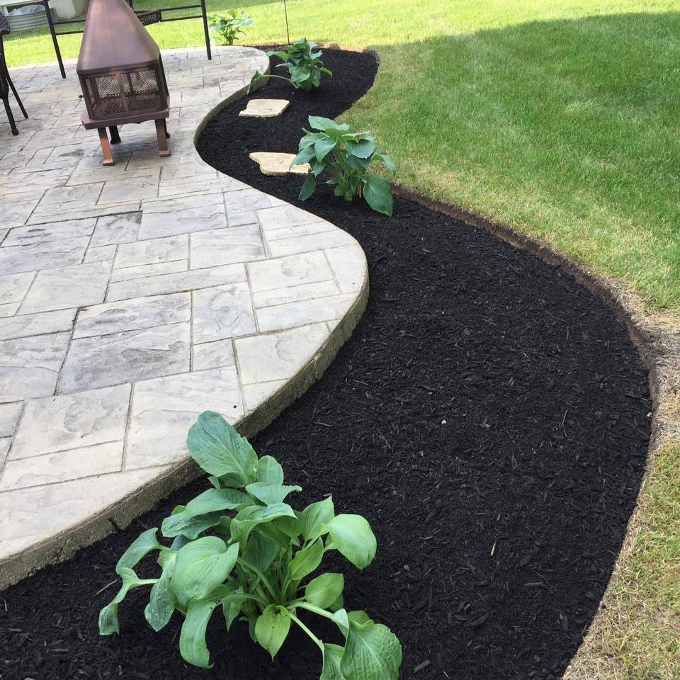 Curved black mulch bed with green plants borders a patio and lawn.