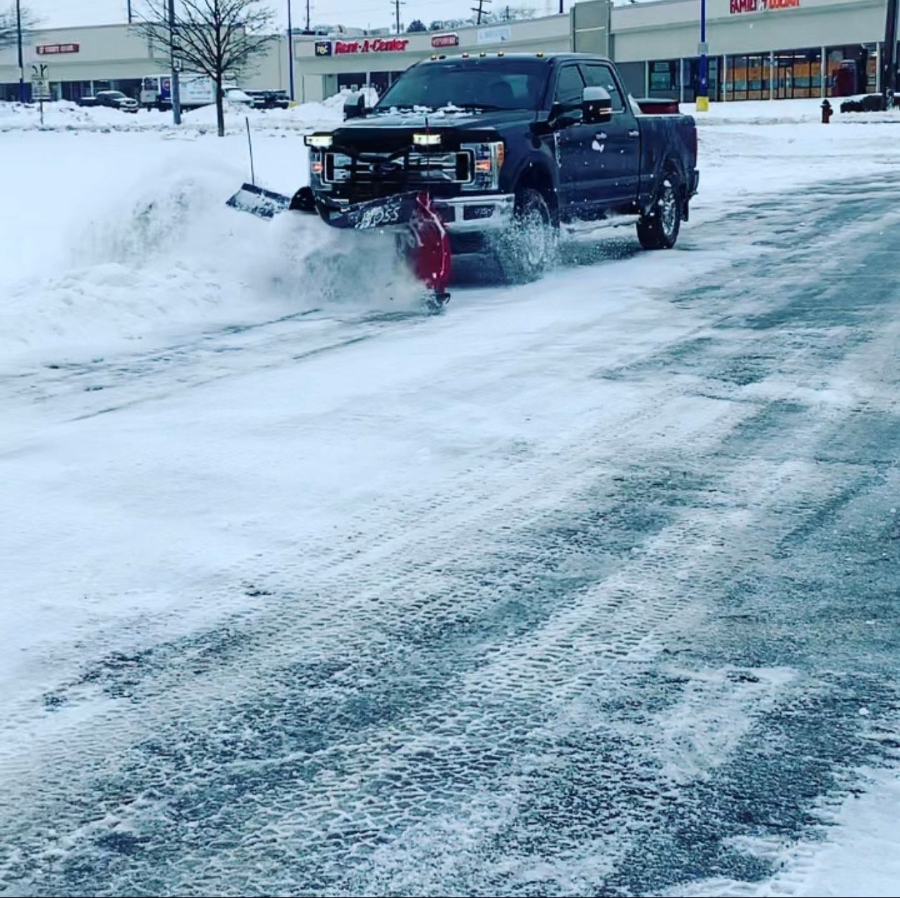 Snow plow truck clearing a snow-covered road in front of businesses on a snowy day.