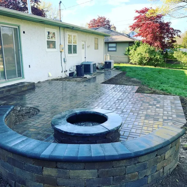 Backyard patio with fire pit, pavers, and retaining wall. House in the background with greenery.