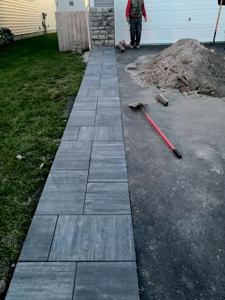 A person stands near a gray paver walkway under construction next to a driveway and lawn.