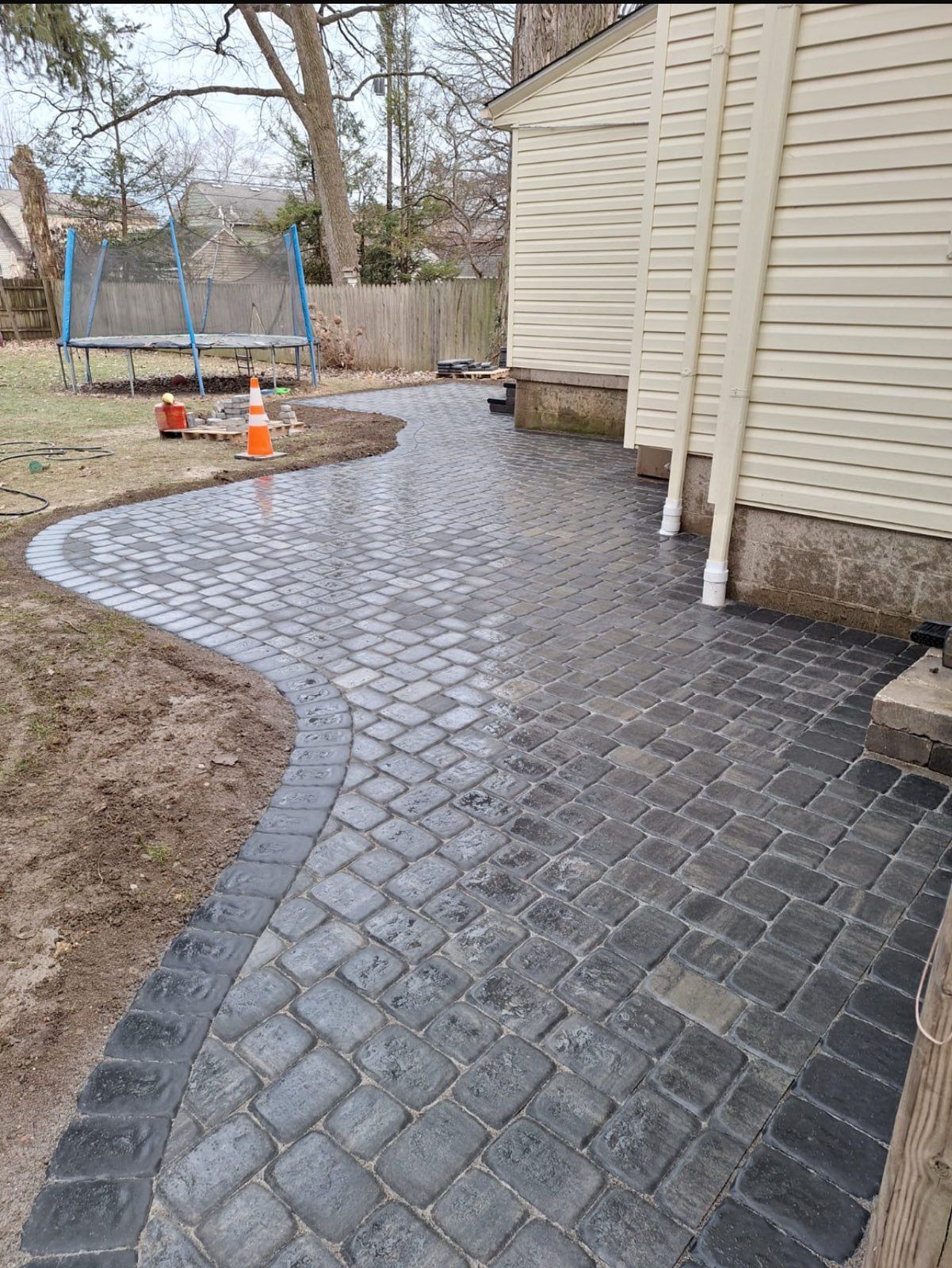 Brick pathway alongside a beige building and lawn with a trampoline in the background.