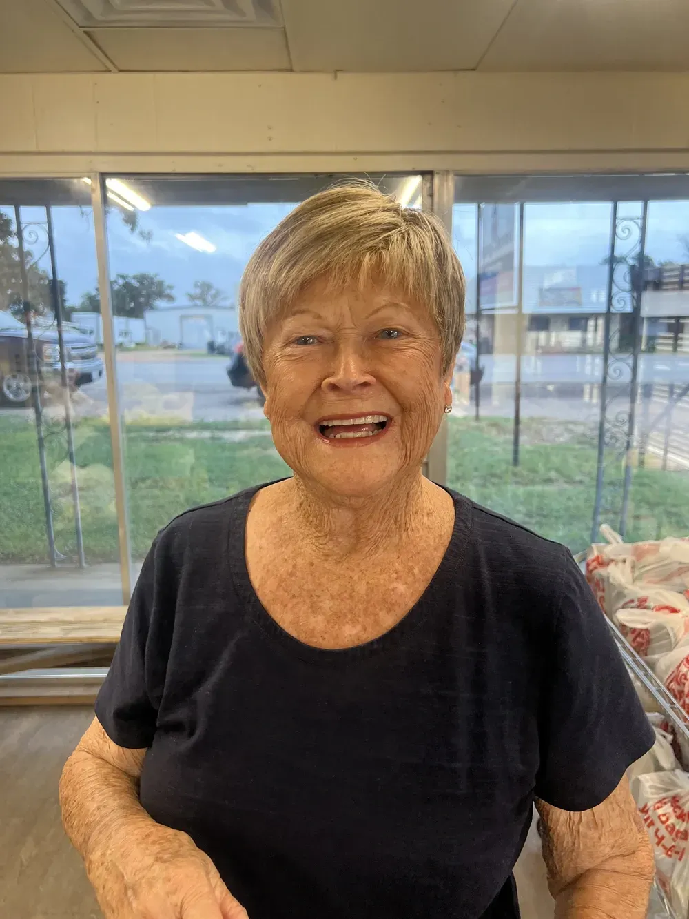 An elderly woman is smiling in a room in front of a window.