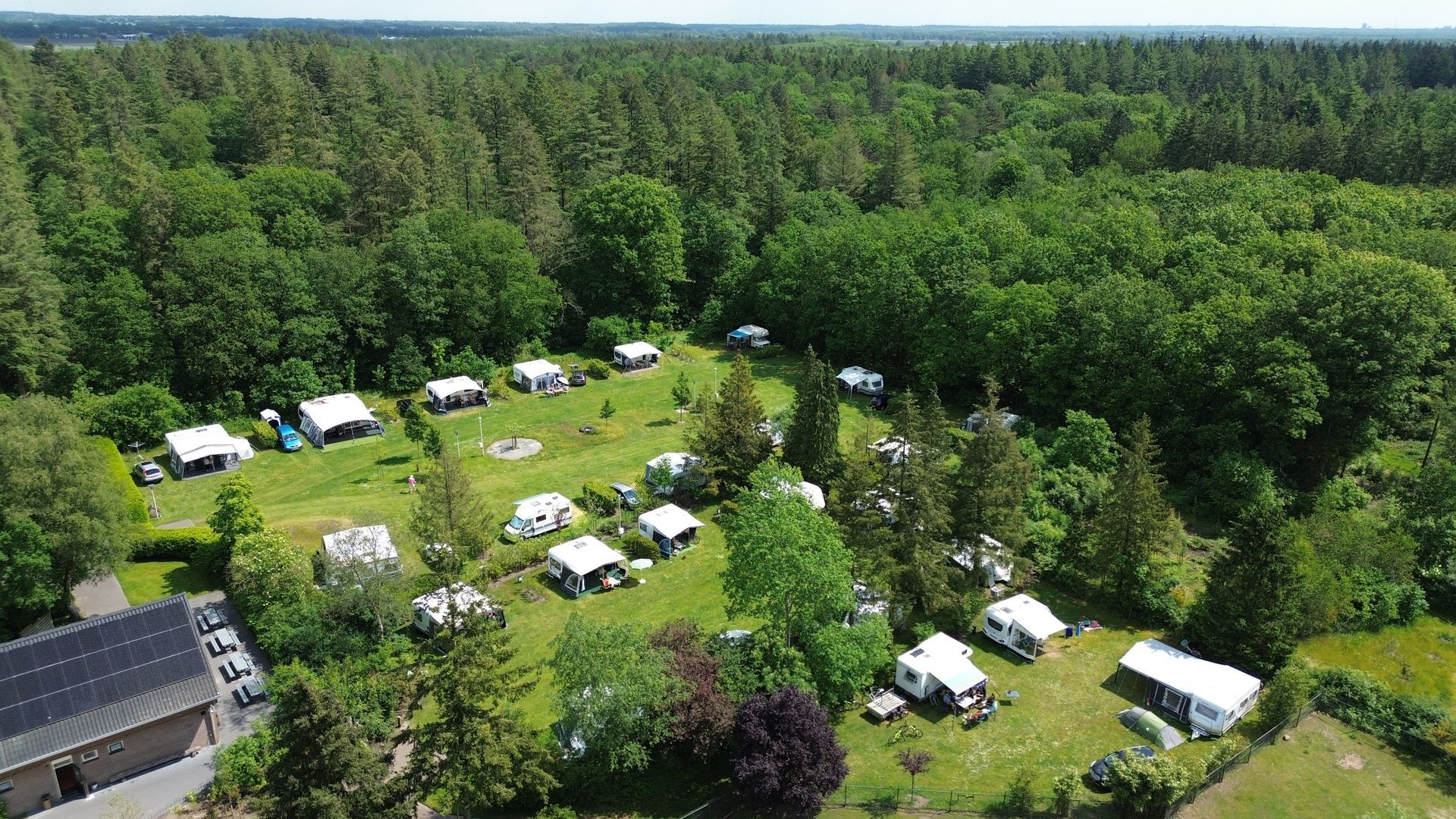 Luchtfoto van een camping met caravans en tenten op een grasveld, omgeven door een weelderig groen bos.
