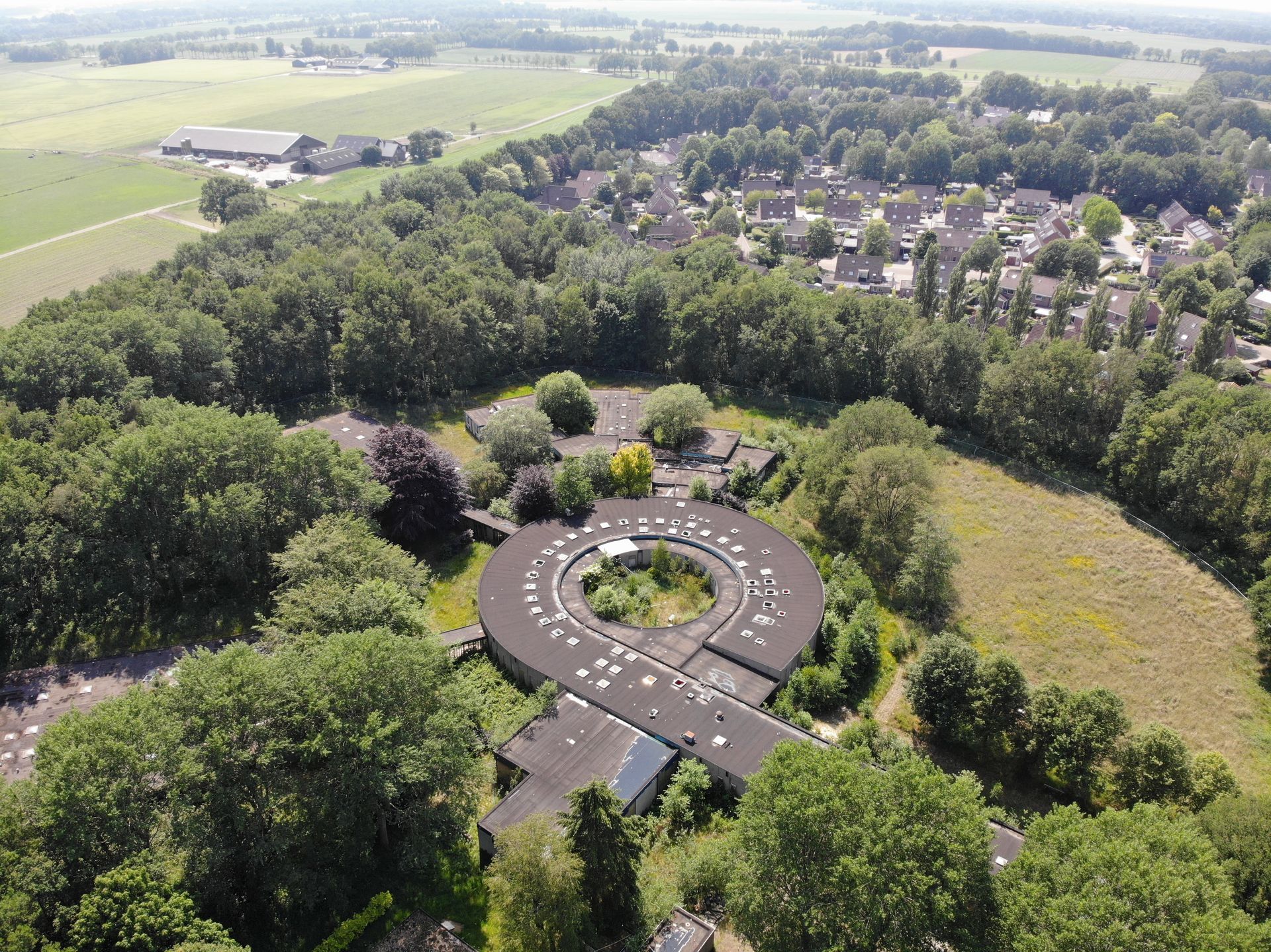 Luchtfoto van een rond, laag gebouw omgeven door bomen, met een stad en velden in de verte.