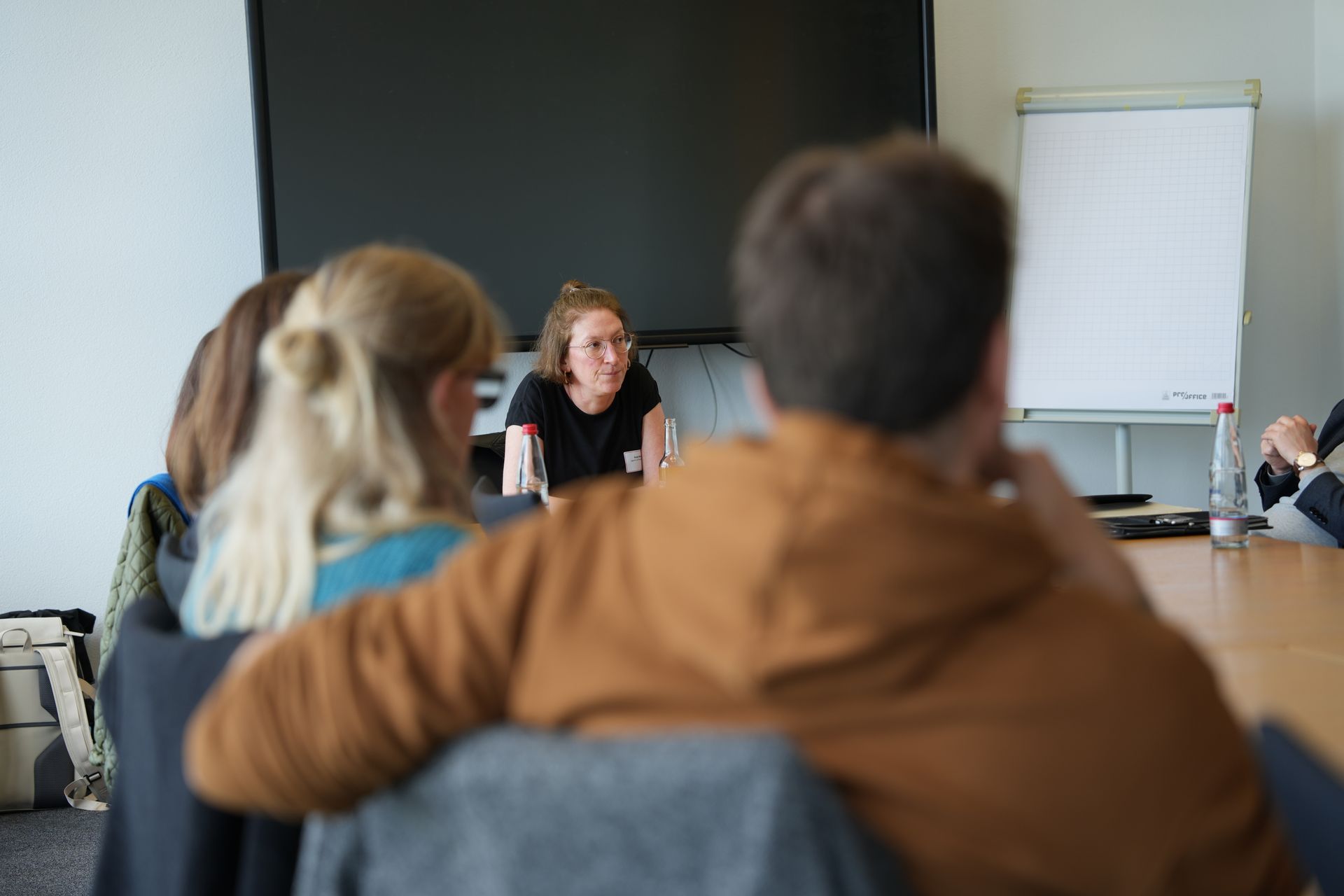 Mensen zitten aan een tafel en luisteren naar een spreker voor een scherm en een whiteboard.