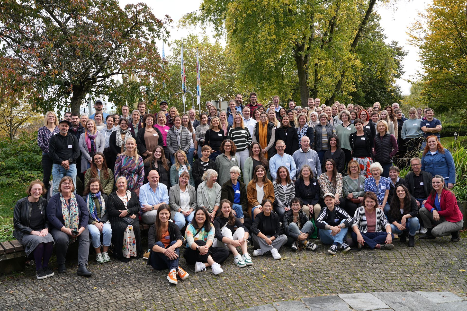 Een grote groep mensen poseert buiten op een bewolkte dag, sommigen lachend, voor een parkachtige omgeving.