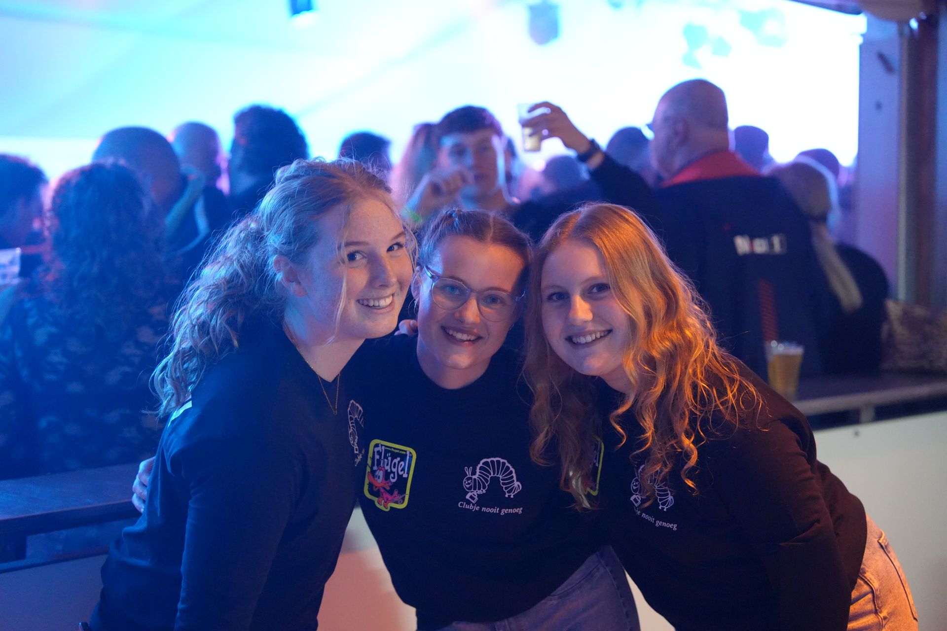 Drie lachende mensen in donkere shirts poseren voor een foto tijdens een evenement, met mensen op de achtergrond.