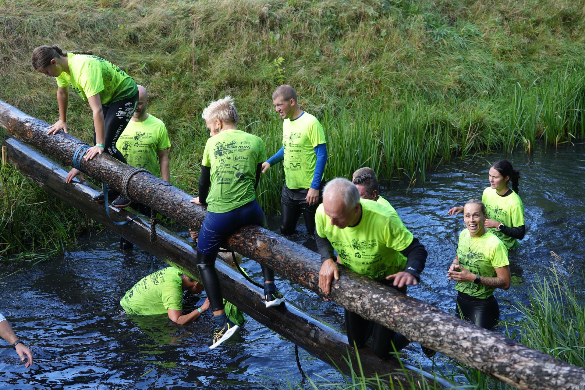 Mensen in felgroene shirts lopen over een houten brug over een beek.