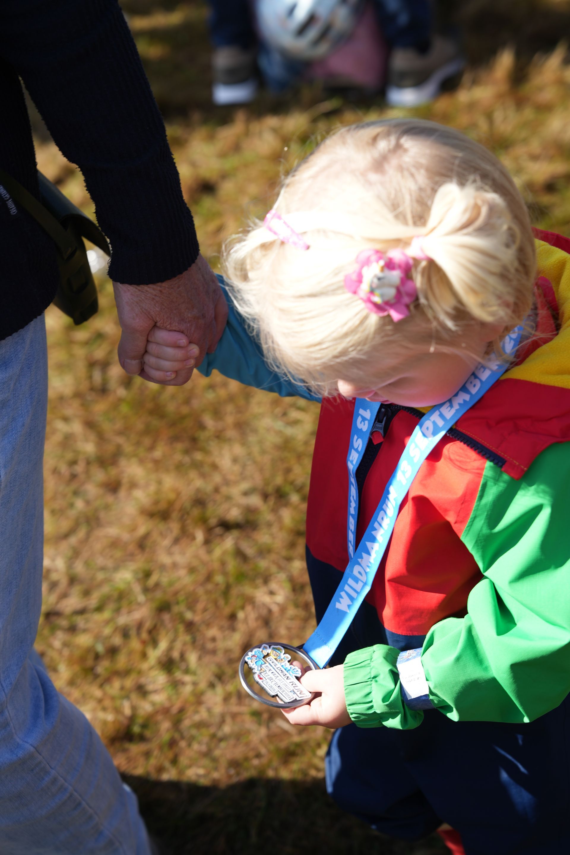 Kind met blond haar kijkt naar een medaille en houdt de hand vast van iemand in een spijkerbroek.