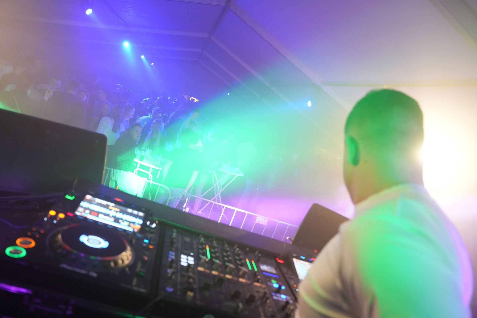Een dj draait muziek op een evenement en mixt achter een mengpaneel. Felle podiumverlichting verlicht een dansende menigte.