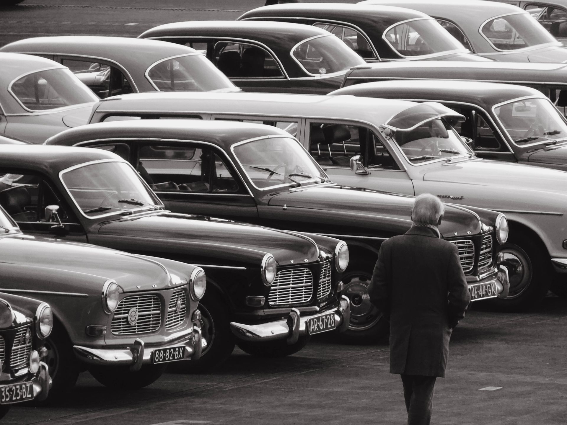 Man loopt langs een rij klassieke auto's die buiten geparkeerd staan. Zwart-witfoto.