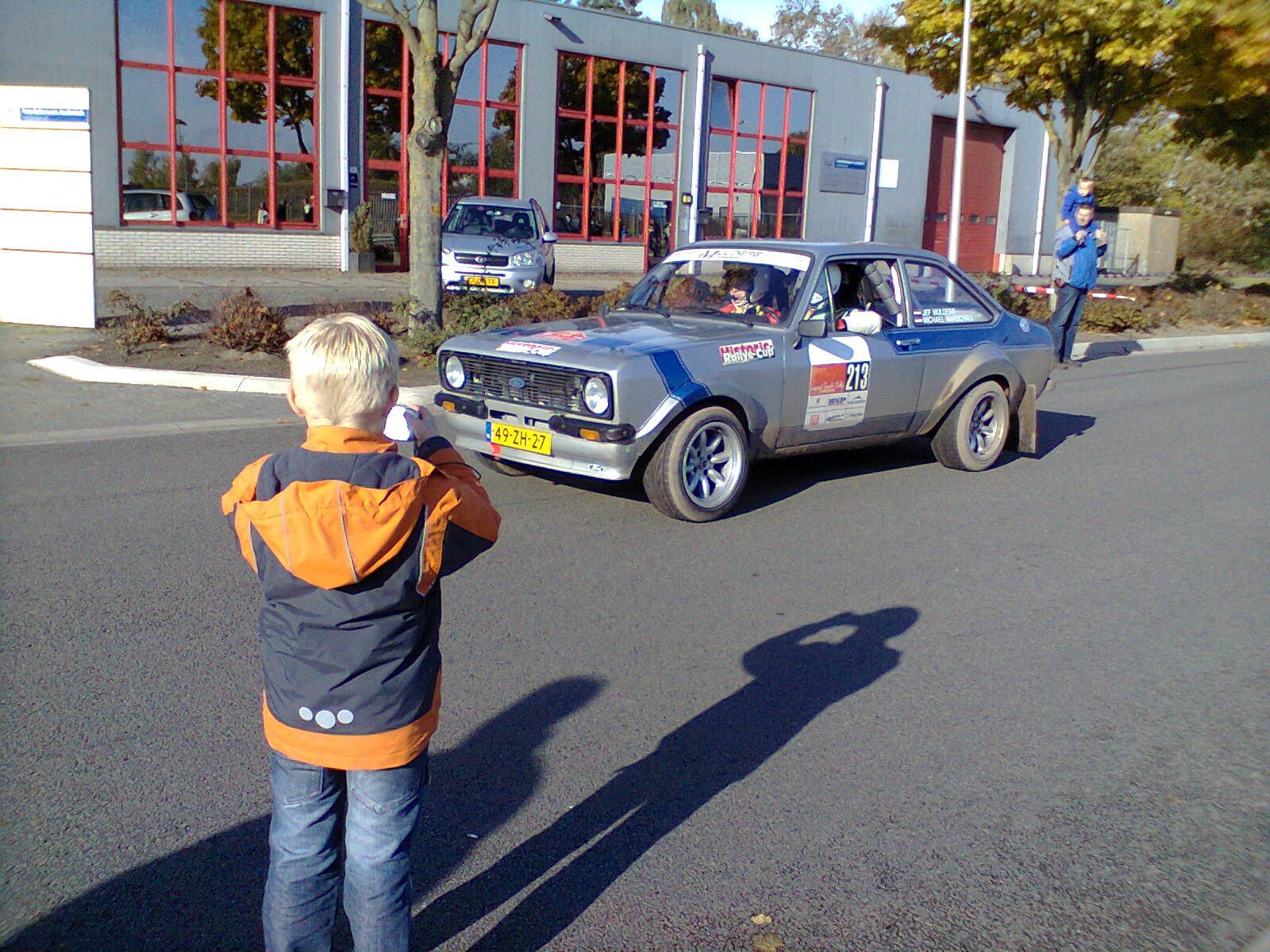 Een jong persoon staat op de stoep, tegenover een rallyauto met blauwe en grijze accenten, voor een gebouw.