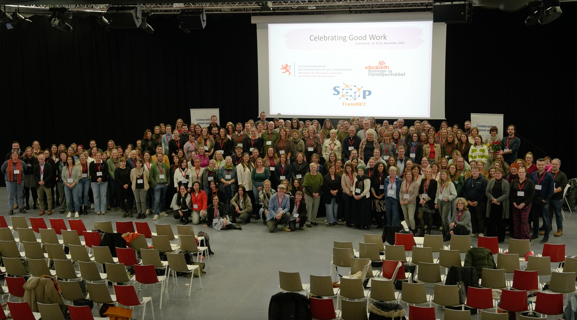 Een groep mensen op een conferentie poseert voor een scherm waarop logo's worden weergegeven. Lege stoelen op de voorgrond.