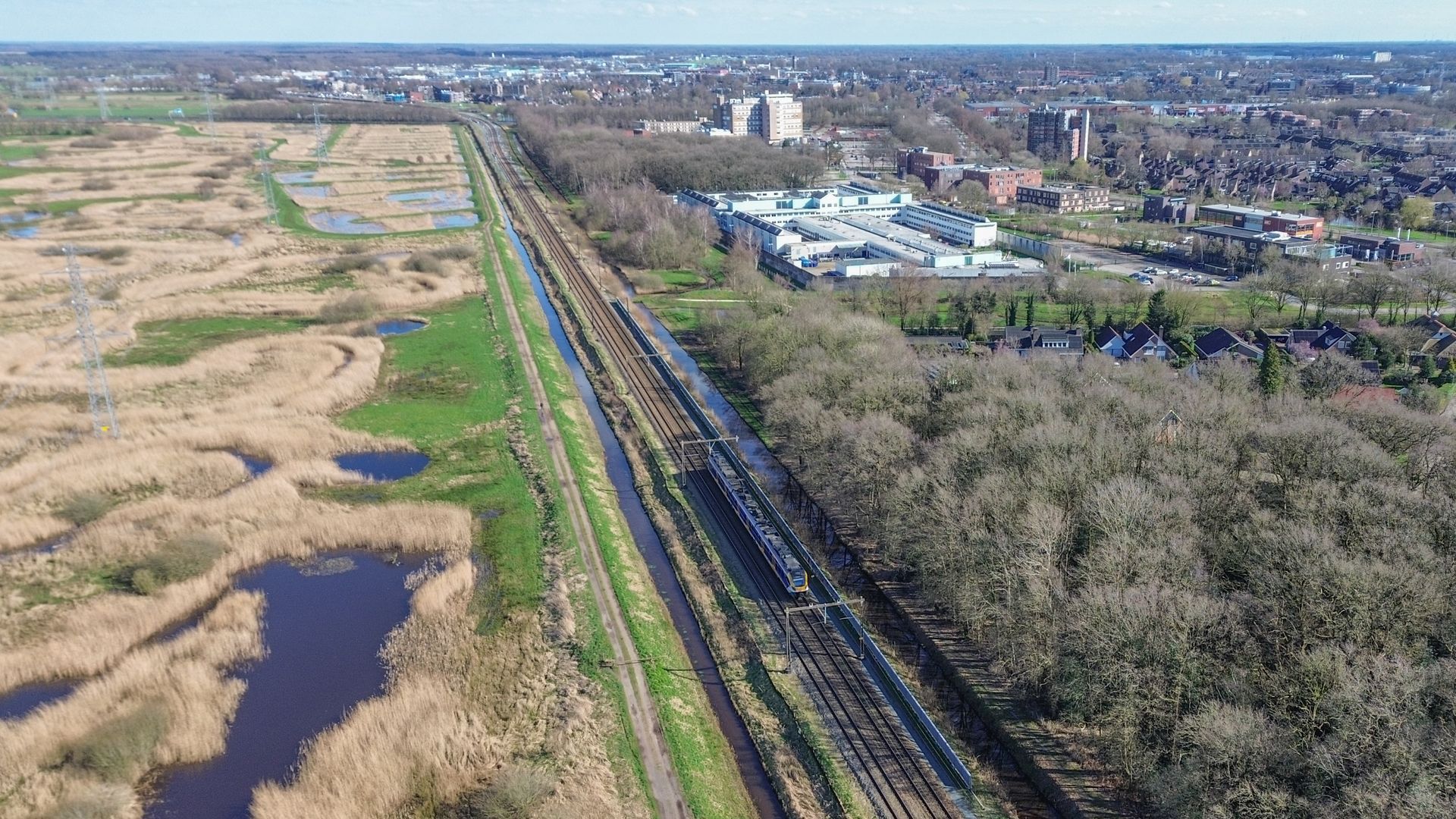 De trein rijdt door een landschap met aan de ene kant moerasgebieden en aan de andere kant een bosrijk gebied met gebouwen.