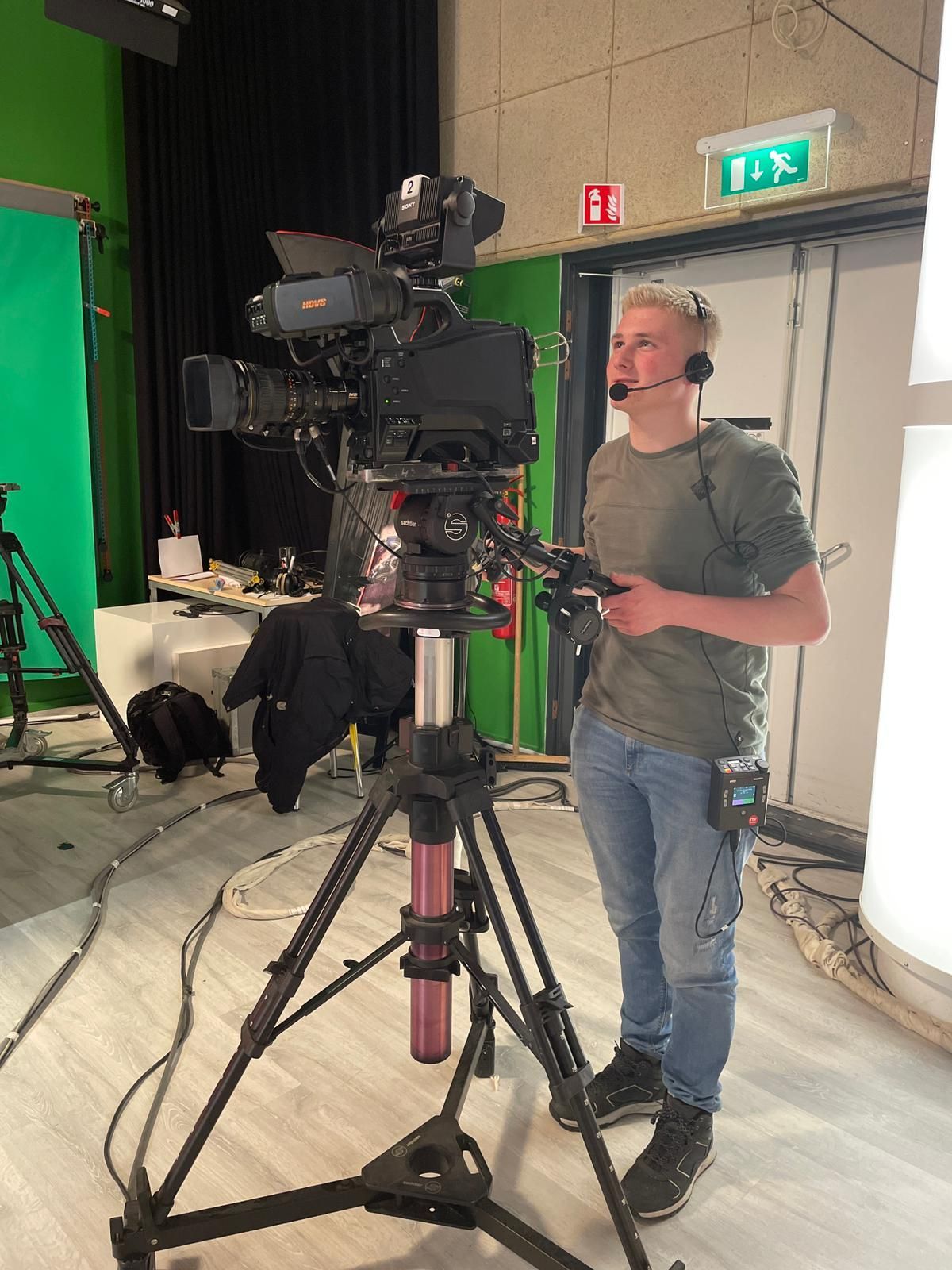 Een man met een headset bedient een televisiecamera op een statief in een studio met een groen scherm.
