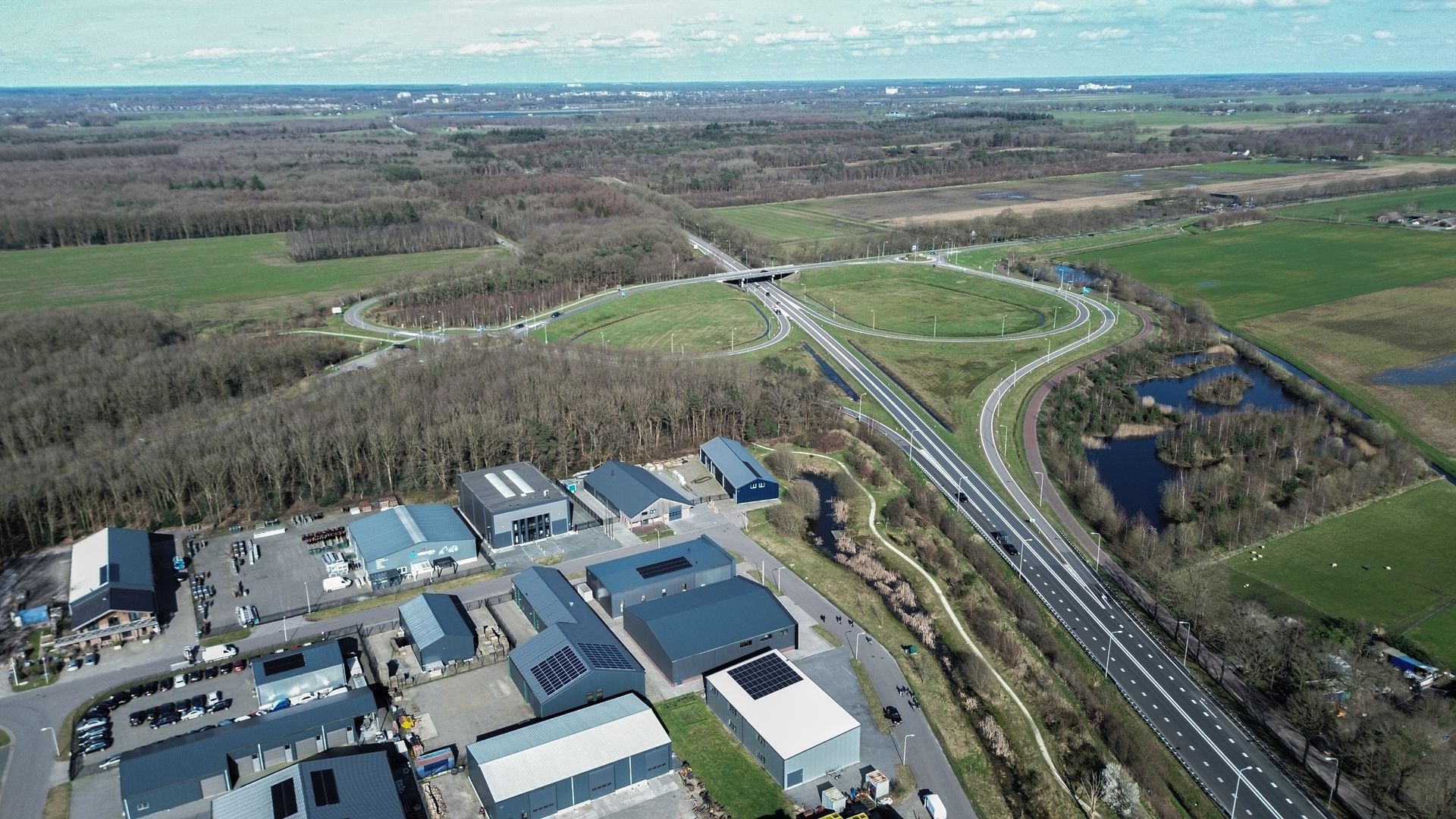 Luchtfoto van een snelwegknooppunt met klaverbladopritten, omgeven door velden en een bedrijventerrein.
