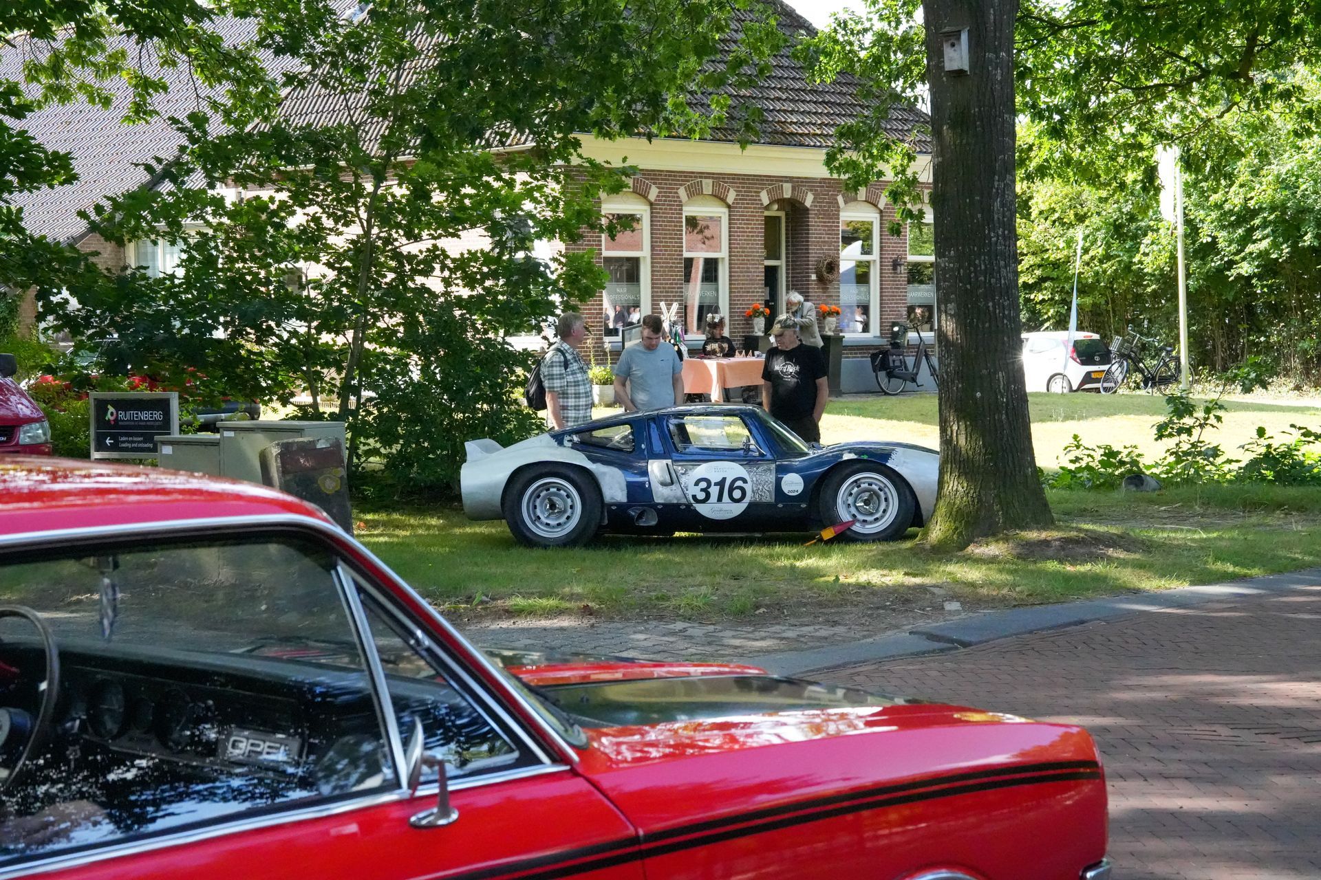 Rode auto op de voorgrond, oldtimer raceauto, mensen bij een gebouw. Groene bomen, zonnige dag.