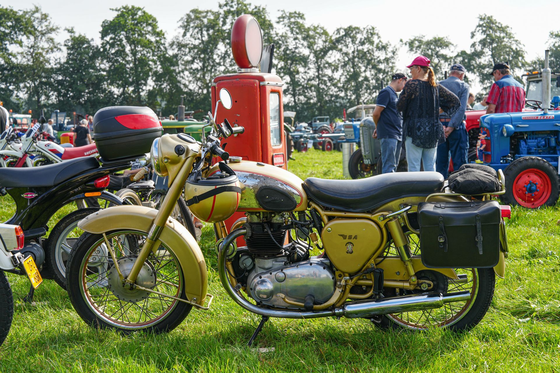 Gele motorfiets met zwarte zadeltassen op een buitenevenement, met mensen en een vintage benzinepomp op de achtergrond.