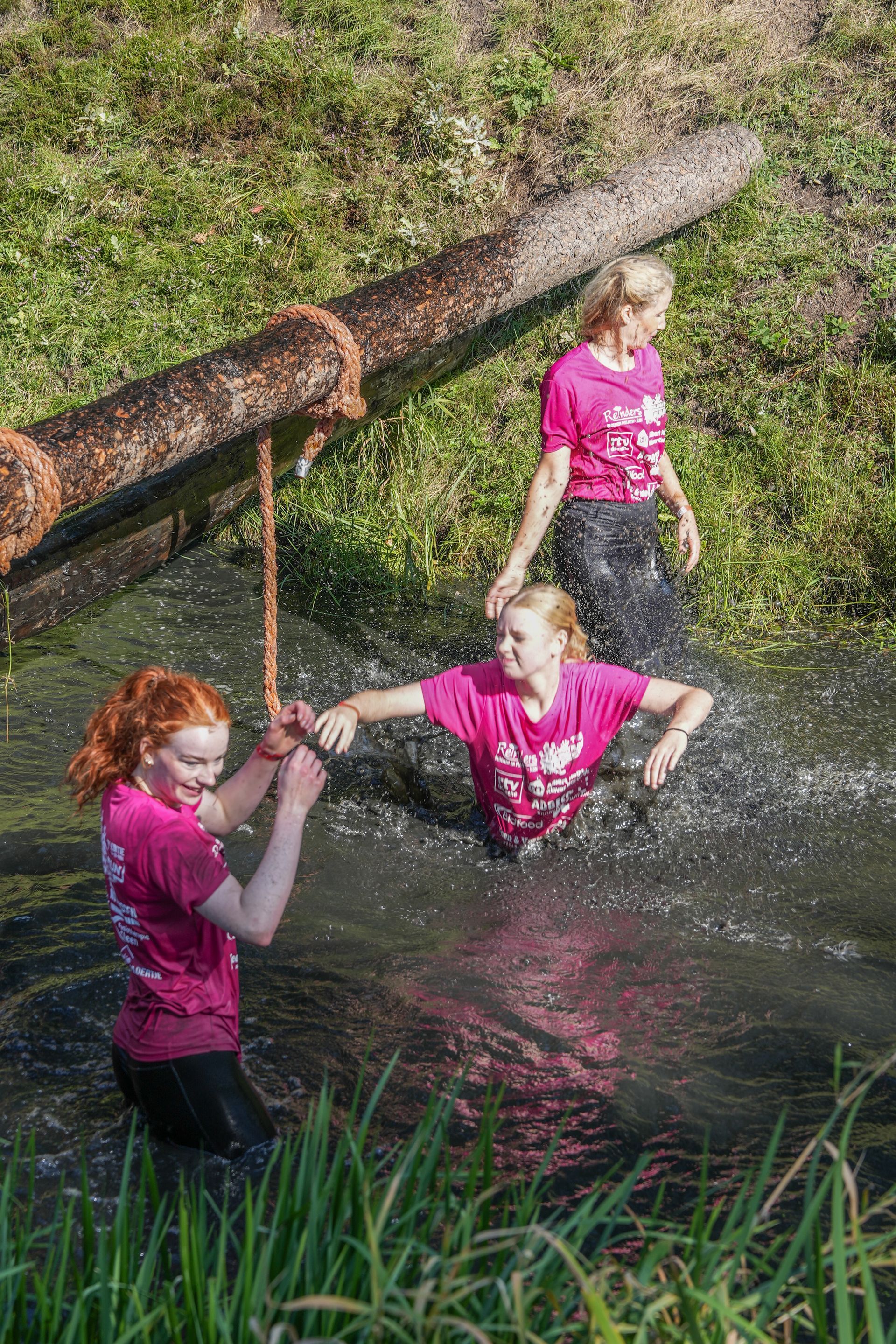 Drie mensen in roze shirts steken een modderige beek over, waarbij ze zich vastgrijpen aan een touw en een boomstam.