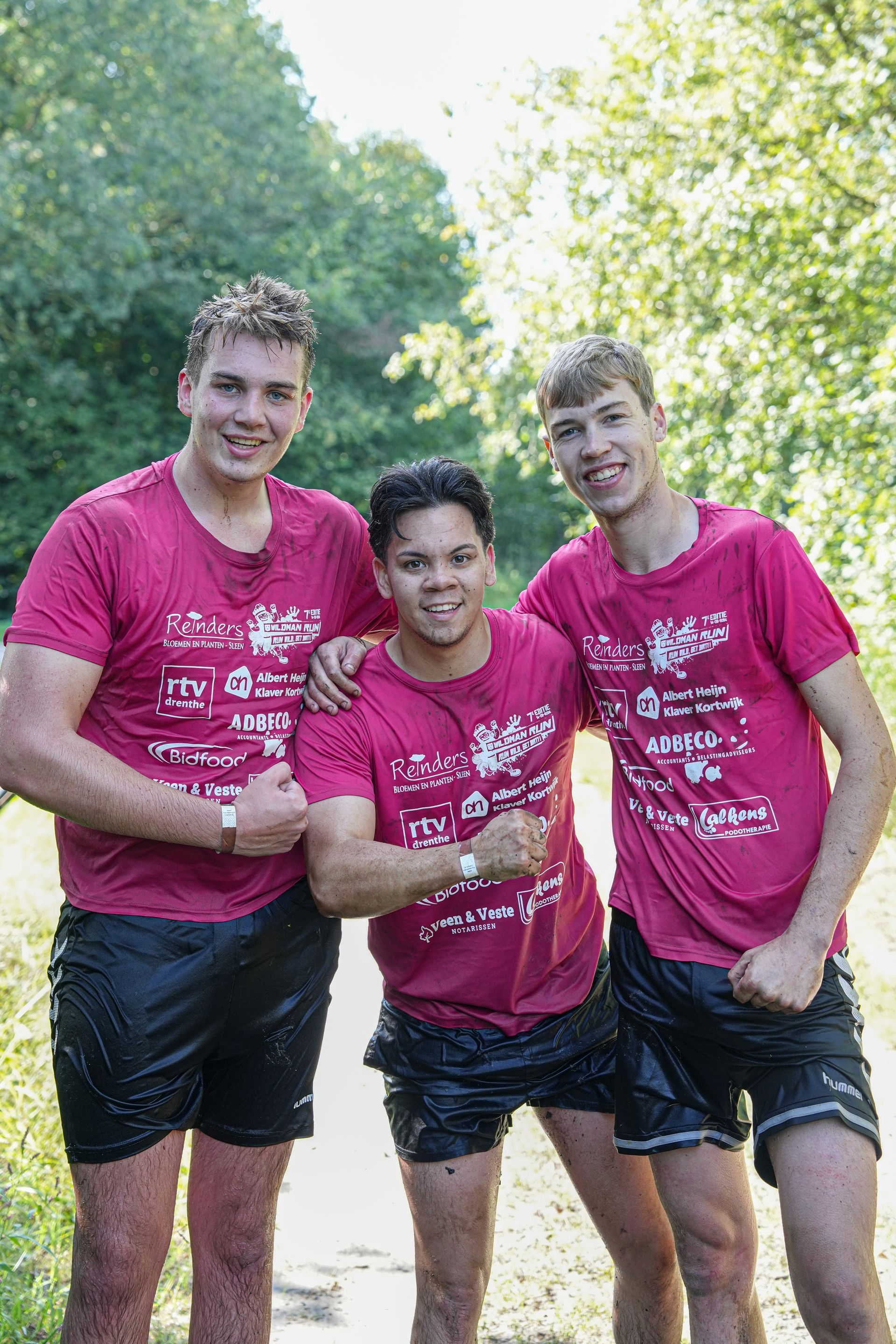 Drie mensen in natte roze shirts en zwarte shorts laten hun spieren zien in een bosrijk gebied.