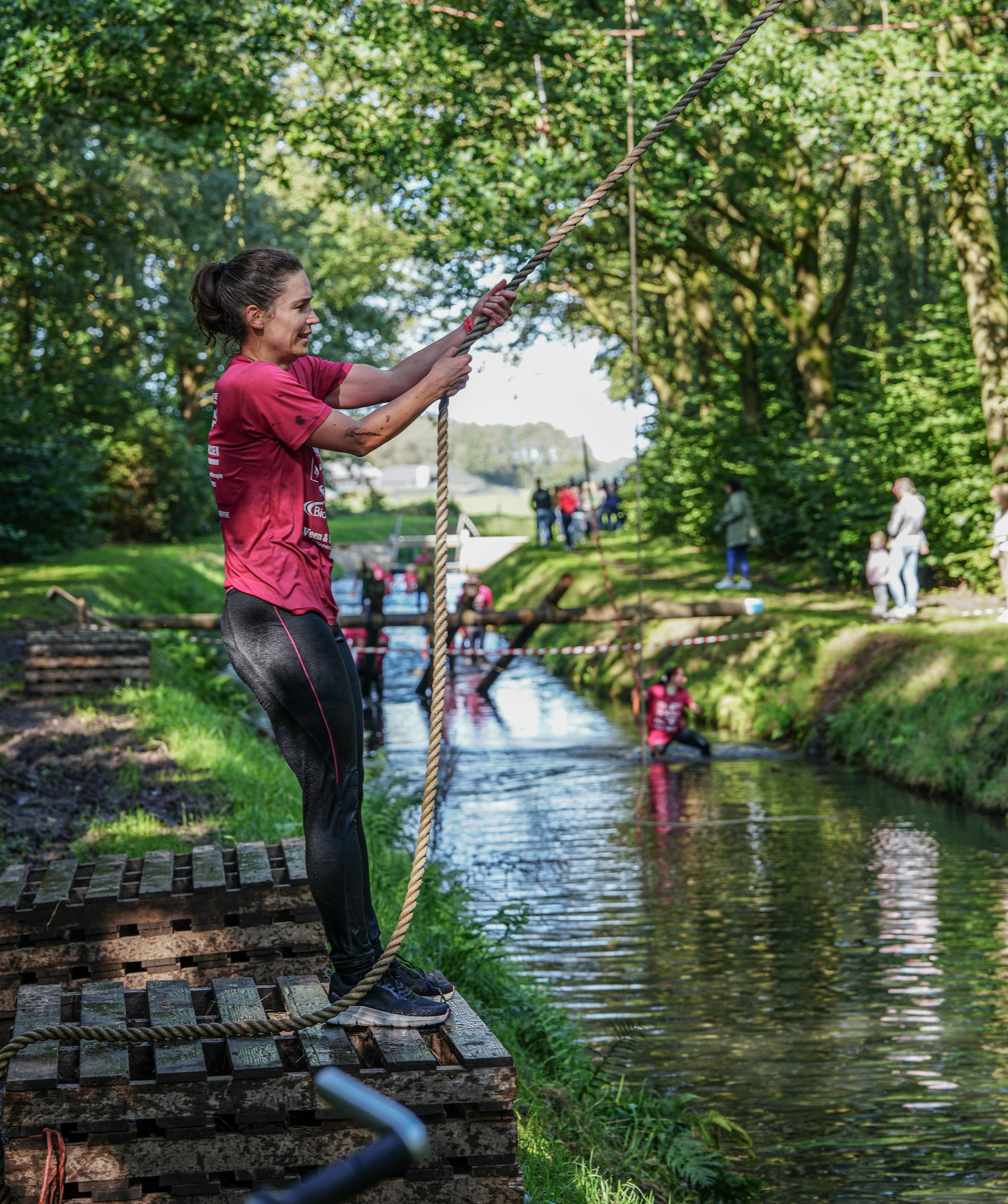 Vrouw op een hindernisbaan, die een touw vastgrijpt, klaar om over het water te slingeren. Parkomgeving met mensen.