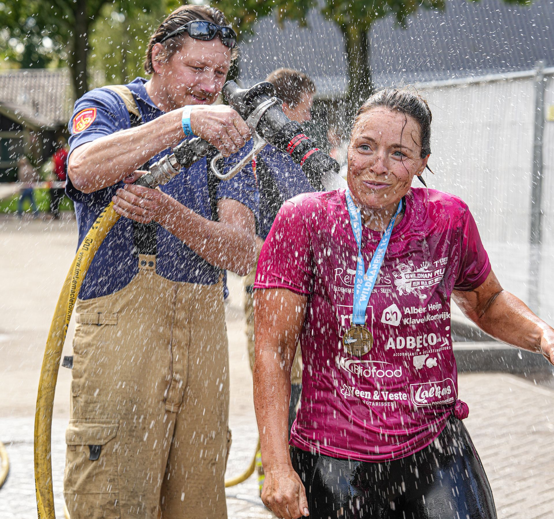 Een brandweerman spuit water op een vrouw met een medaille; buiten, na een wedstrijd.