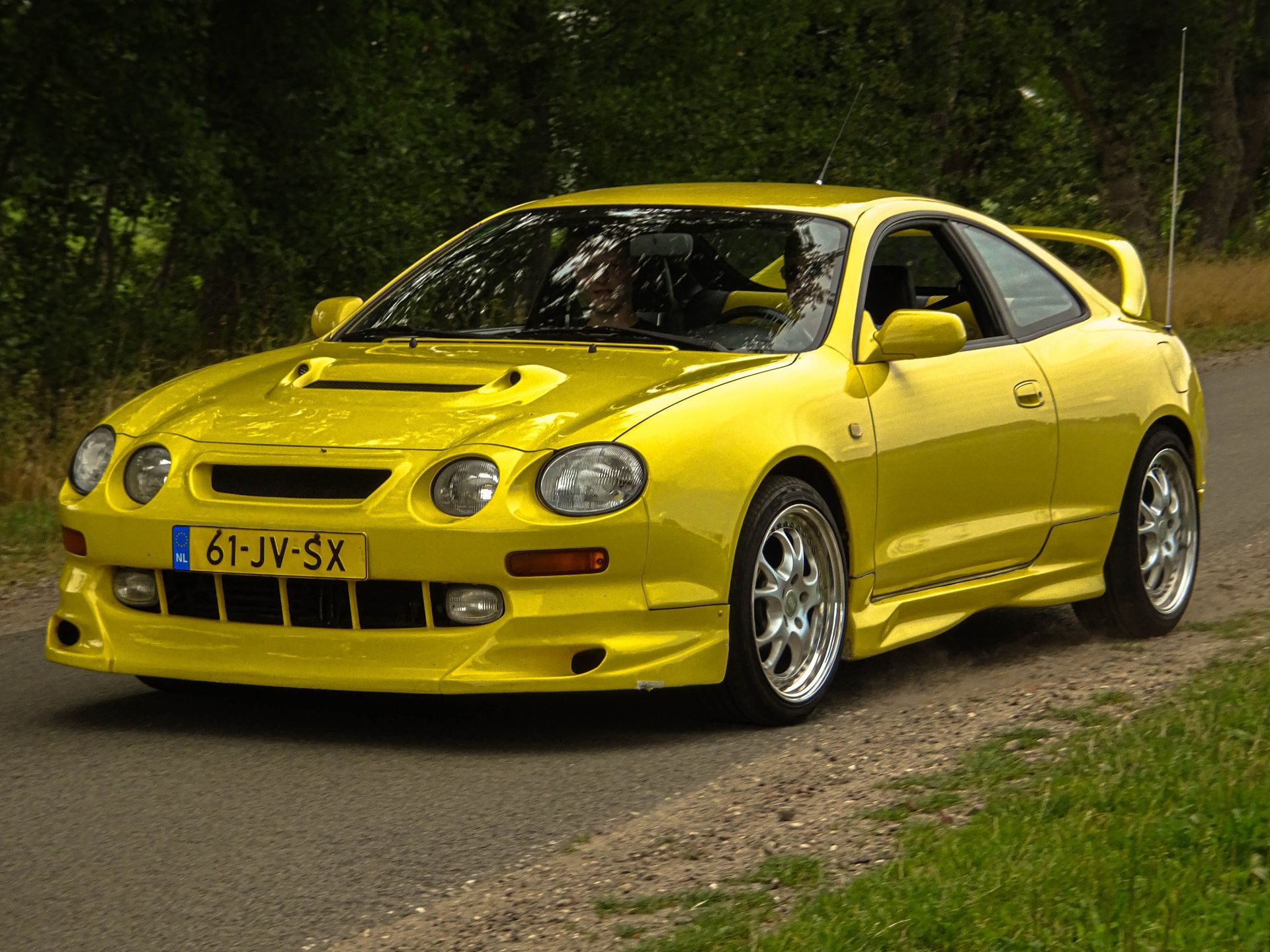 Een gele Toyota Celica coupé met een aangepaste bodykit staat geparkeerd op de weg.