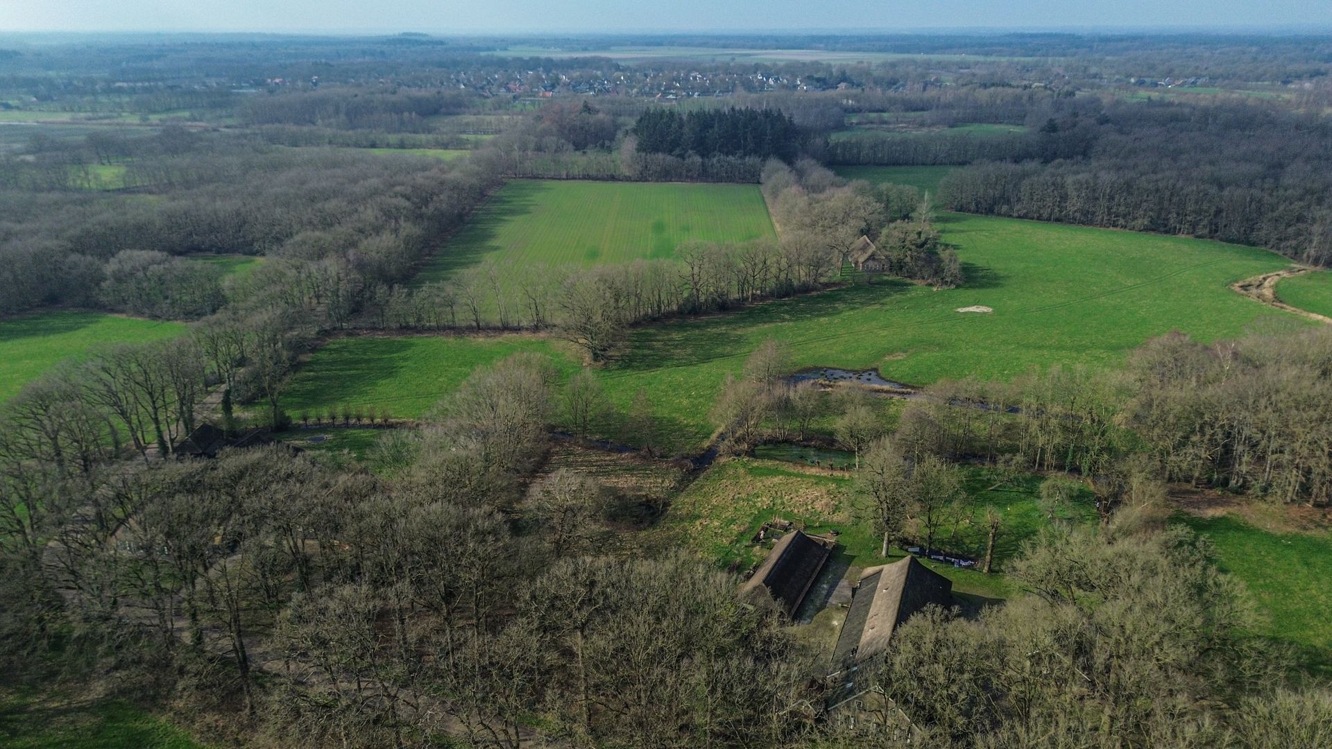 Luchtfoto van een groen veld omgeven door bomen, met een klein gebouw op de voorgrond en een stad in de verte.