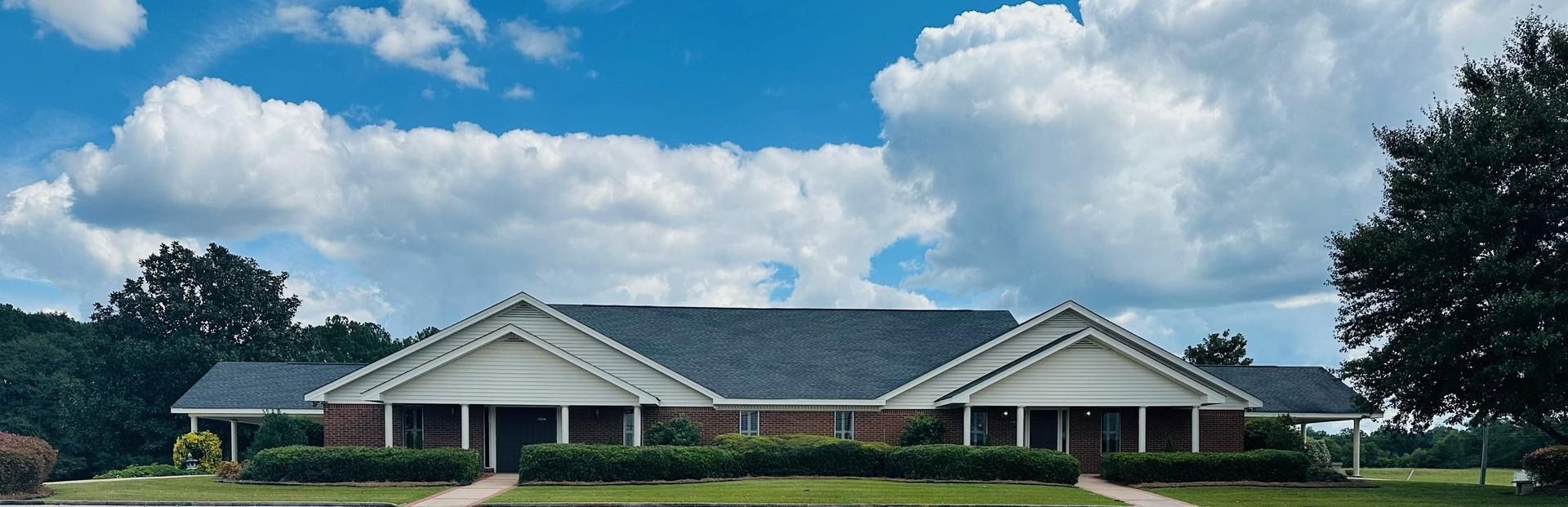 Single-story brick building with white columns and a dark roof under a partly cloudy sky