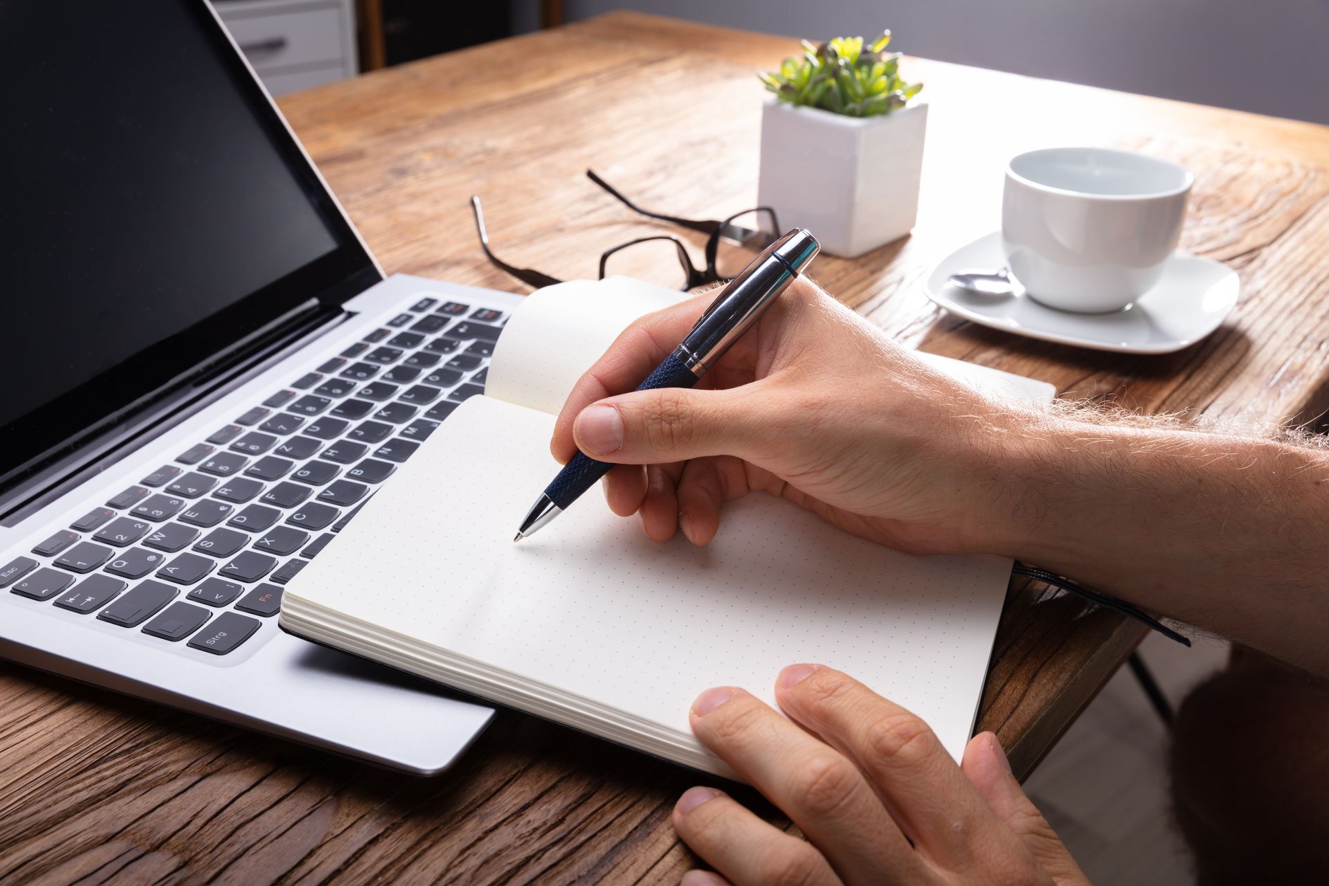 Hand writing in a notebook beside a laptop on a wooden desk with coffee and a small plant.