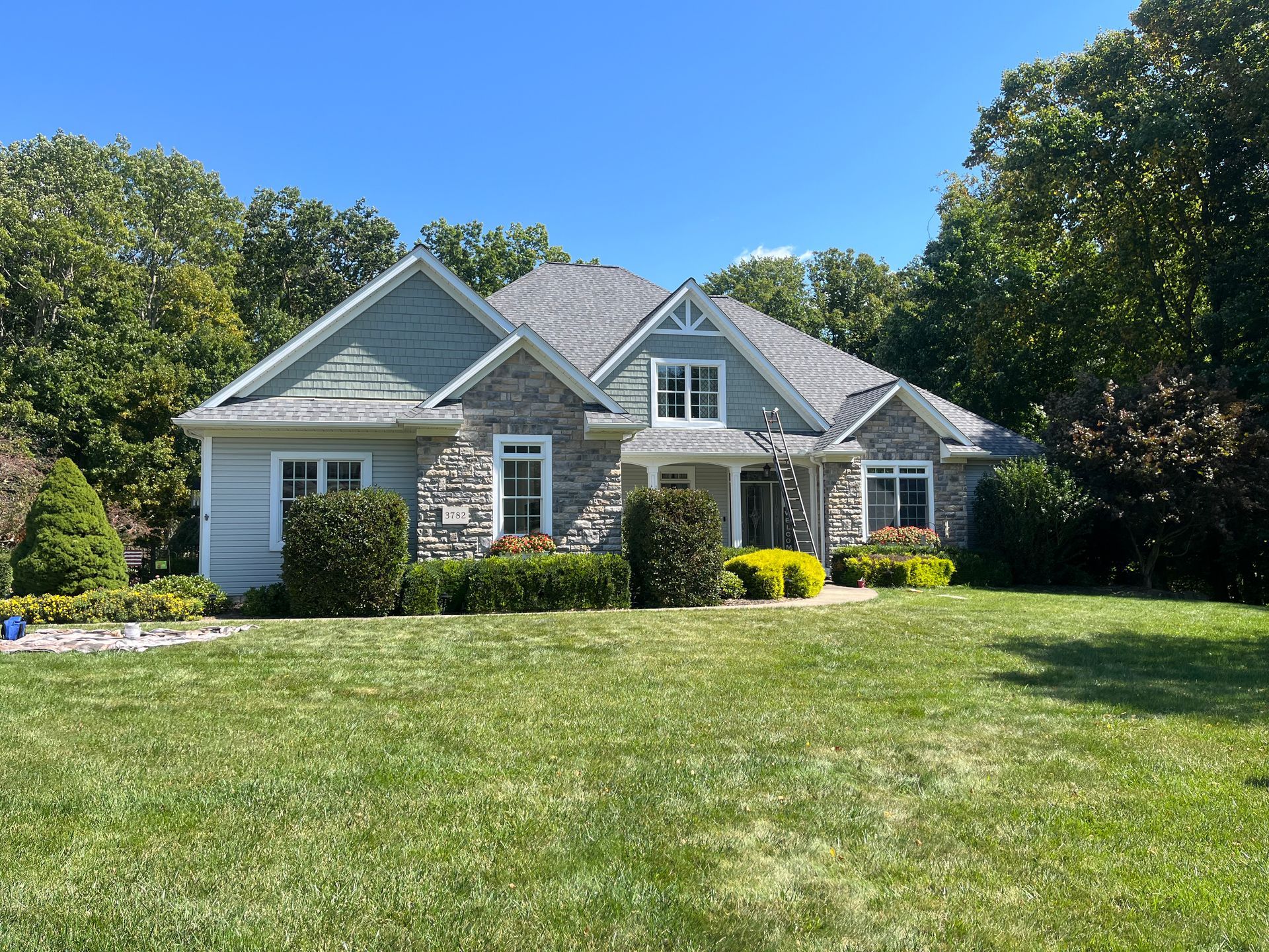House with gray stone and blue-green siding, gray roof, and lush green lawn under a clear blue sky.