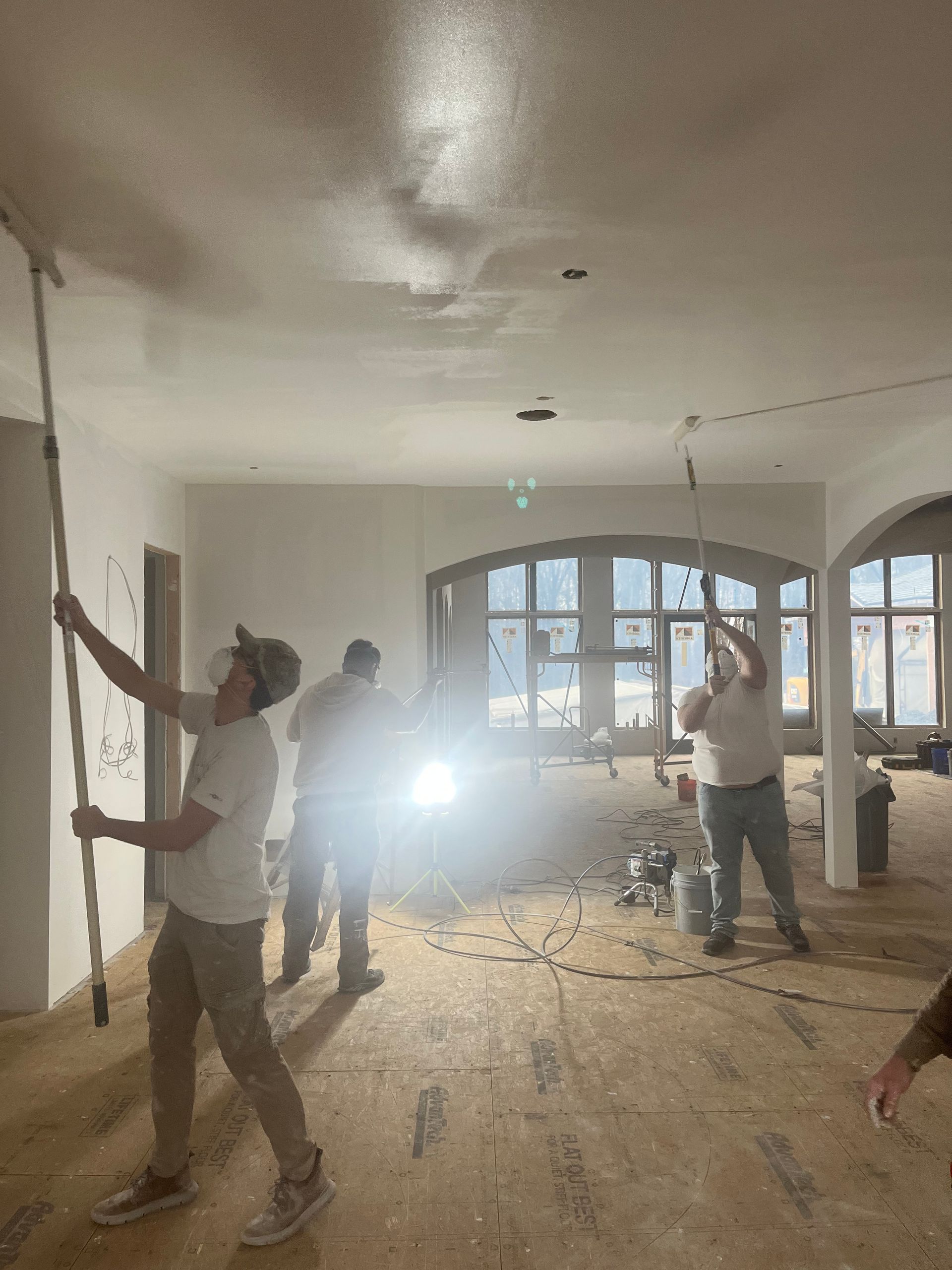 People sanding a ceiling in a room with large windows, light, and drywall dust.
