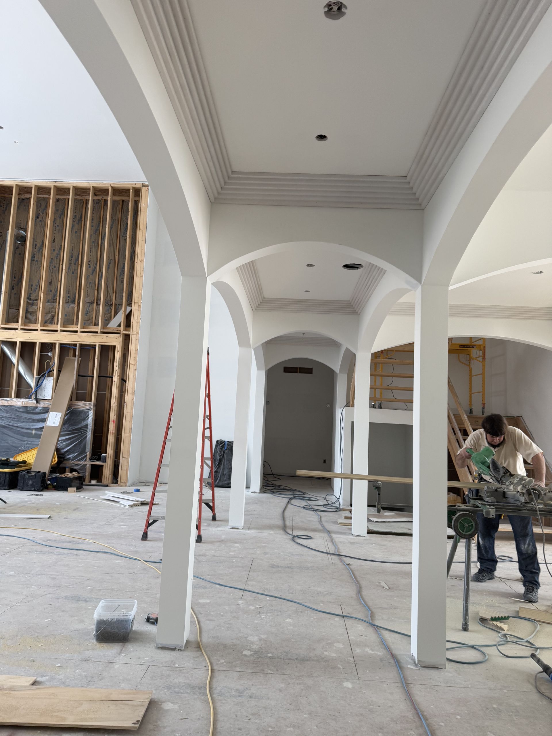 Construction site interior with arched doorways, drywall, and worker using a saw.