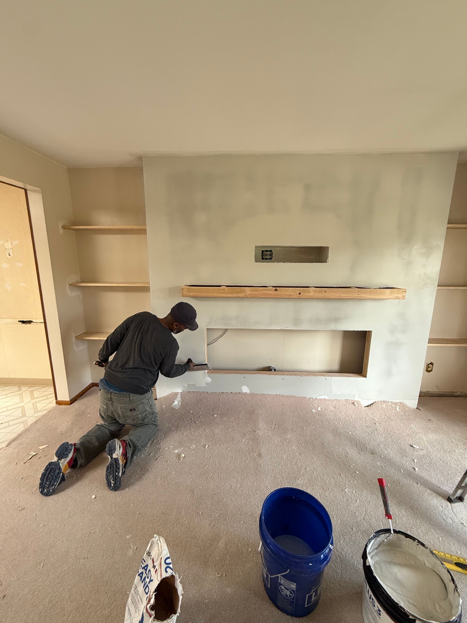 Person applying compound to a fireplace surround. Interior with neutral walls, built-in shelves, and a blue bucket.