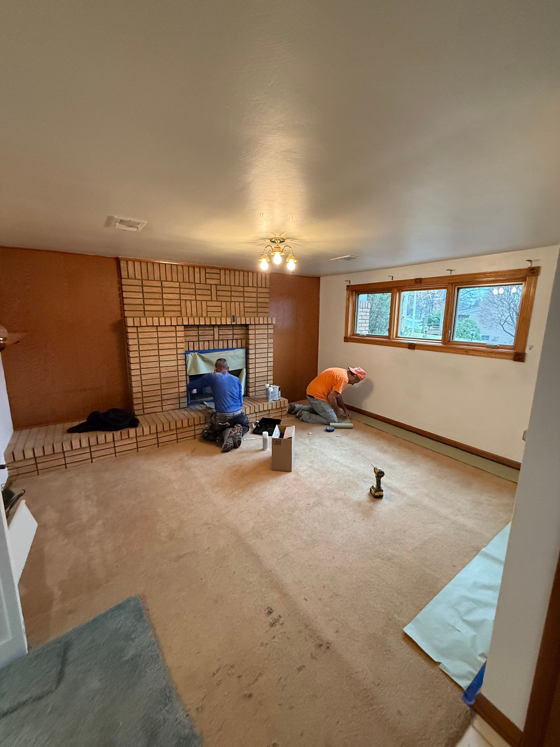 Workers installing flooring in a living room with a brick fireplace and windows; one person is near the wall.