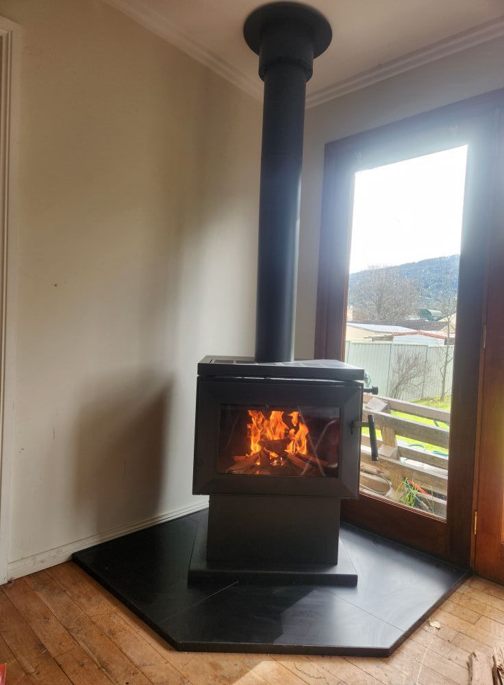Wood-burning stove with lit fire, black metal, installed on a black tile hearth near a sliding glass door.  — Unger Air in South Tamworth, NSW