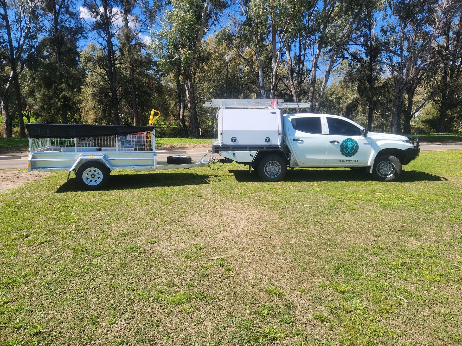A white utility truck with a canopy and a flatbed trailer parked on a grassy field in front of a tree line. — Unger Air in South Tamworth, NSW