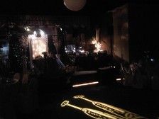 Dark stage lit with spotlights and projector, with a person and instruments visible.
