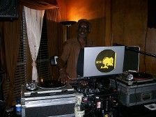DJ behind turntables at a dimly lit venue, wearing brown shirt, logo on laptop screen.